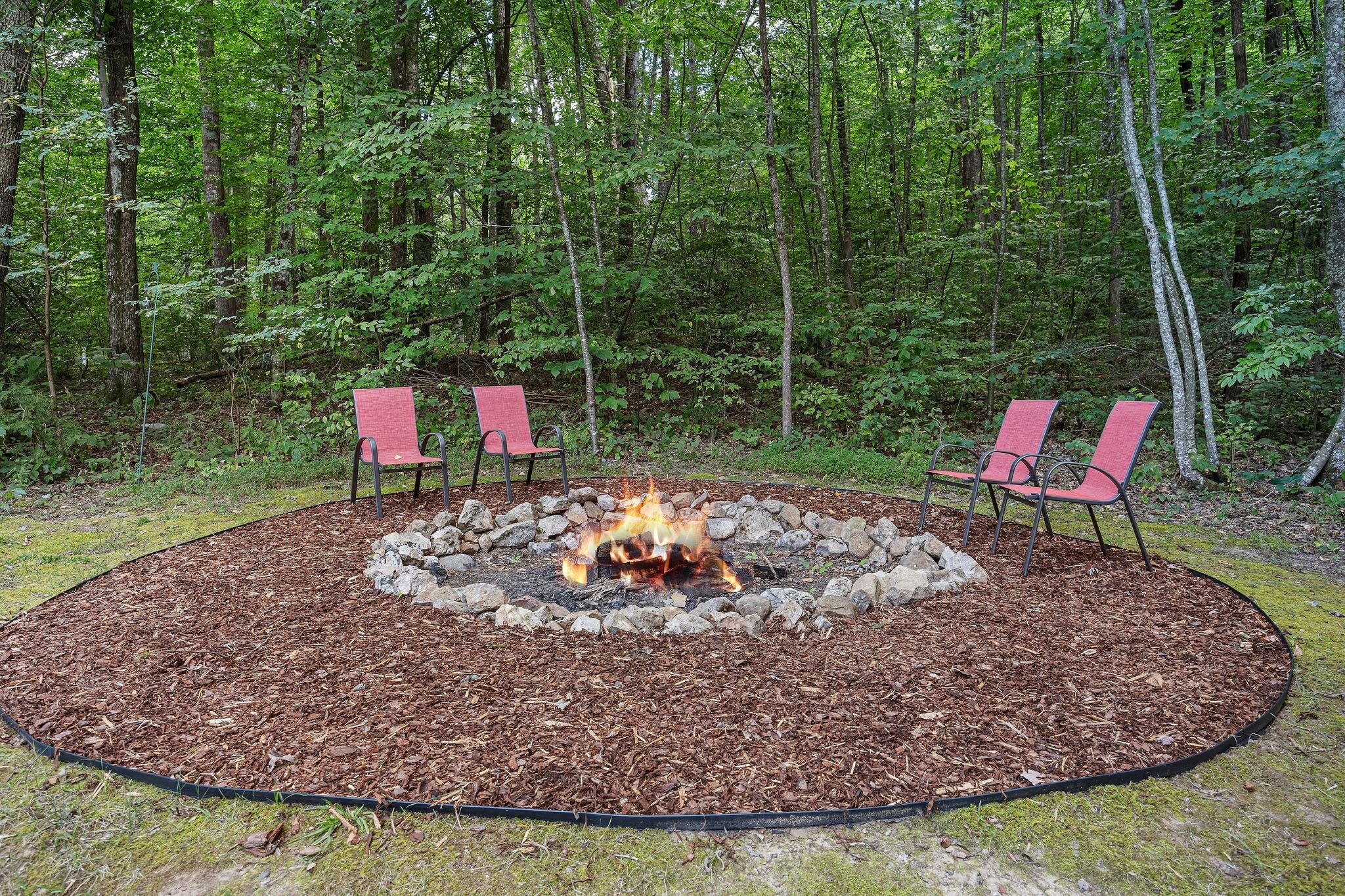 A circular fire pit surrounded by rocks and mulch, with two red chairs facing the fire.