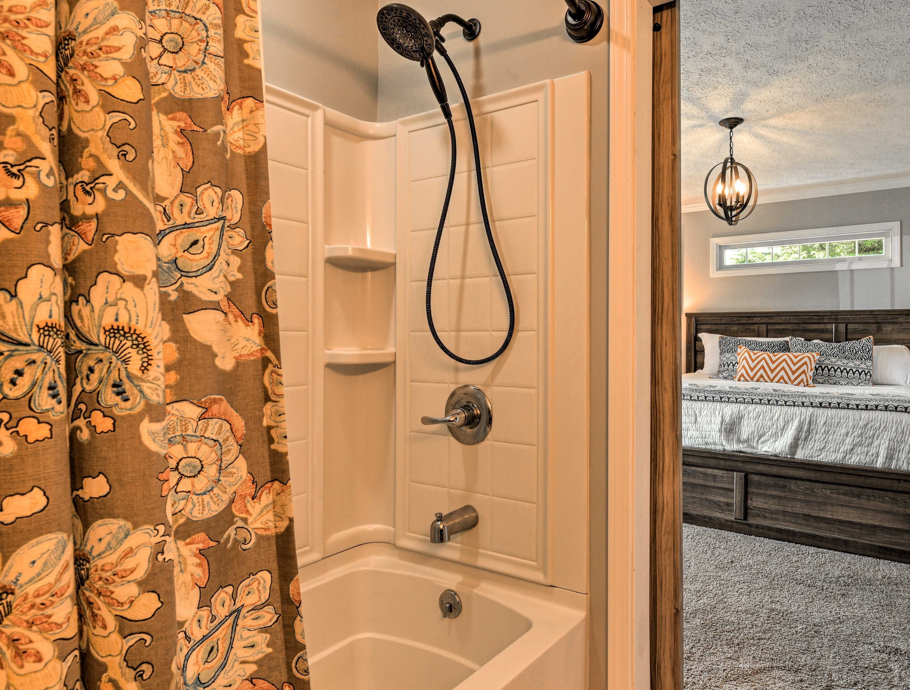 Een badkamer met een douche, een bad en een toilet. In de aangrenzende kamer staat een bed met beddengoed met een patroon.