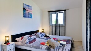 Uma cama de casal com colcha, mesas de cabeceira com luminárias, uma janela com cortinas e um quadro na parede.