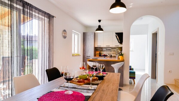 Uma área de refeições com uma mesa de madeira posta para uma refeição, uma cozinha ao fundo e vista para o exterior através de grandes janelas.