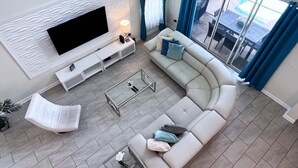 A modern living room with a large flat-screen TV, a white sofa with blue cushions, a white armchair, and a glass coffee table. - 5 Bed 5 Bath South Facing Pool Home Storey Lake 5 Bedroom Home by RedAwning (Kissimmee)