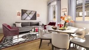 Una sala de estar moderna con un sofá gris, un sillón rojo y una mesa de comedor con sillas blancas. Hay un ventanal con cortinas, un cuadro en la pared y un tapete estampado.
