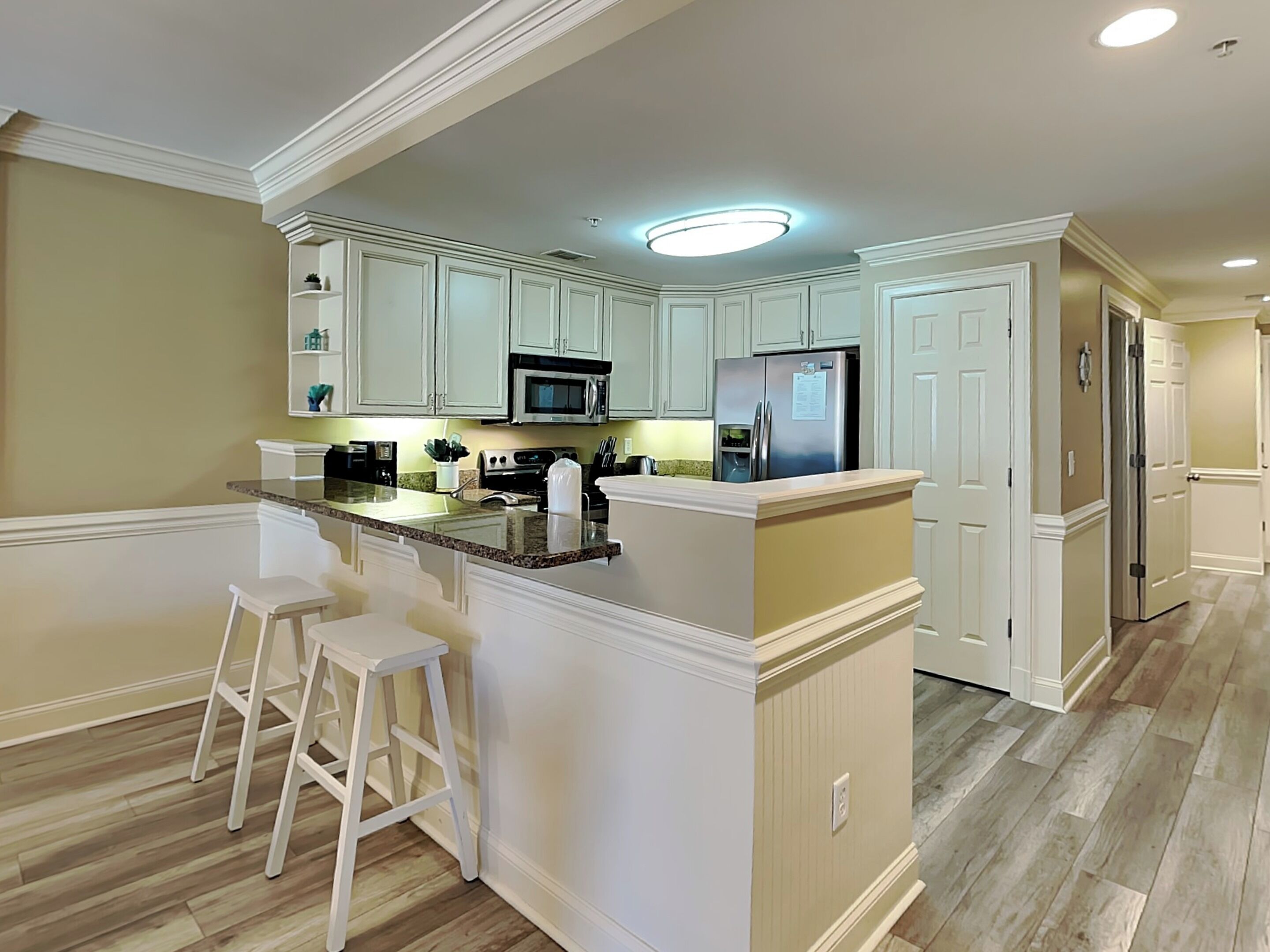 Una cocina con gabinetes blancos, una isla central con taburetes, microondas, refrigerador y horno empotrado.