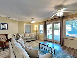 A living room with a sofa, armchair, and coffee table, a ceiling fan, and large windows.