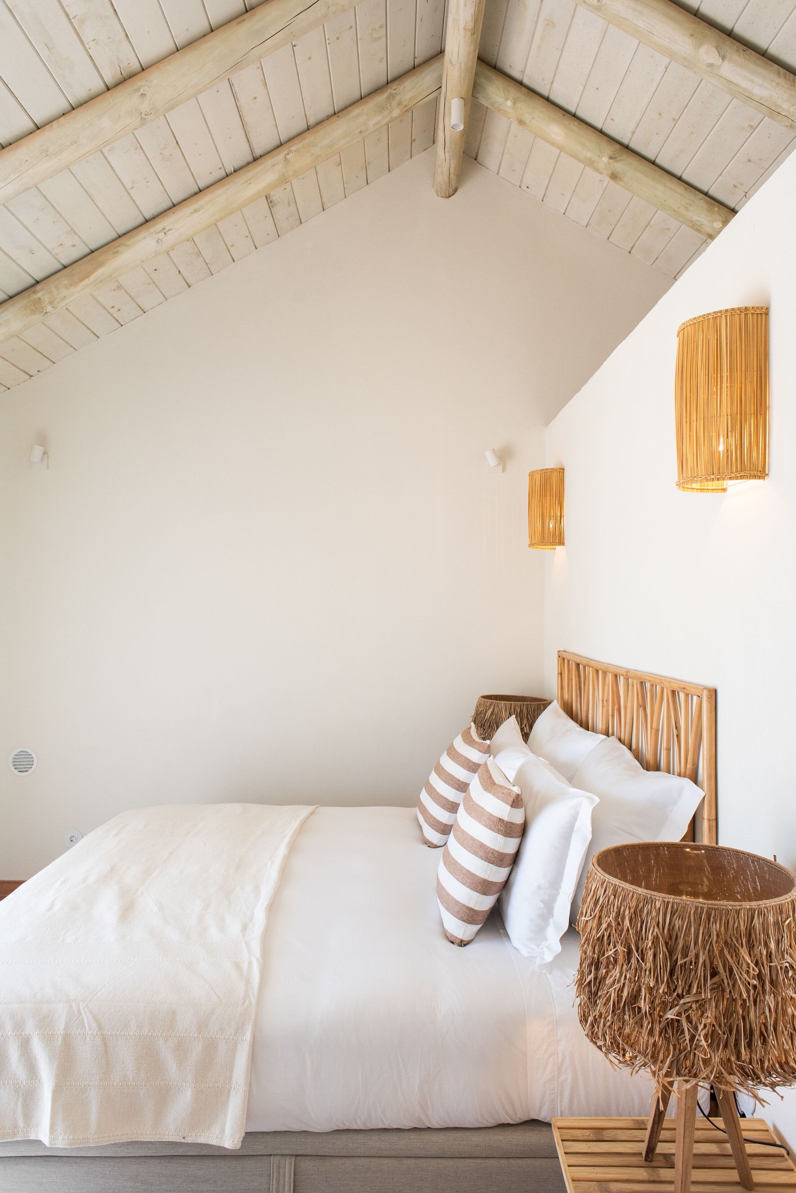 Quarto com teto de madeira, cama com roupa de cama branca, almofadas listradas e mesa de cabeceira de fibra natural.