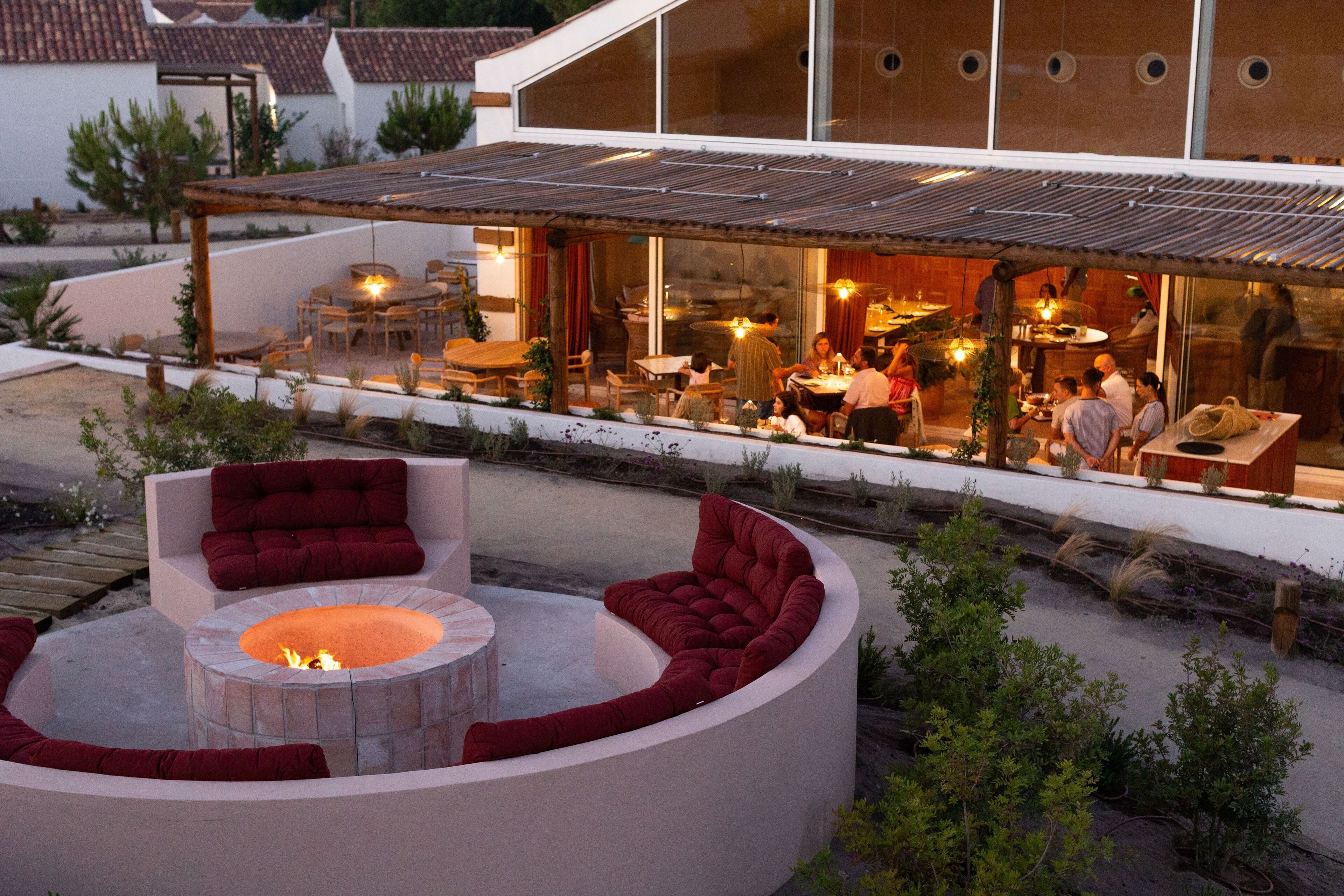 Un área de estar moderna al aire libre con una chimenea, rodeada de sillones y sofás con cojines rojos, con vista a un comedor con comensales.