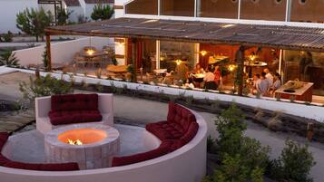 Un área de estar moderna al aire libre con una chimenea, rodeada de sillones y sofás con cojines rojos, con vista a un comedor con comensales.