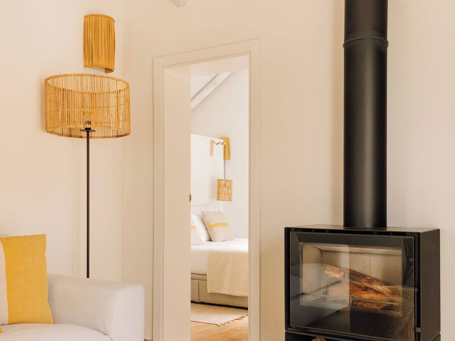 Une pièce chaleureuse avec un canapé blanc, un pouf en fibres tressées, un poêle à bois noir et une lampe en osier.