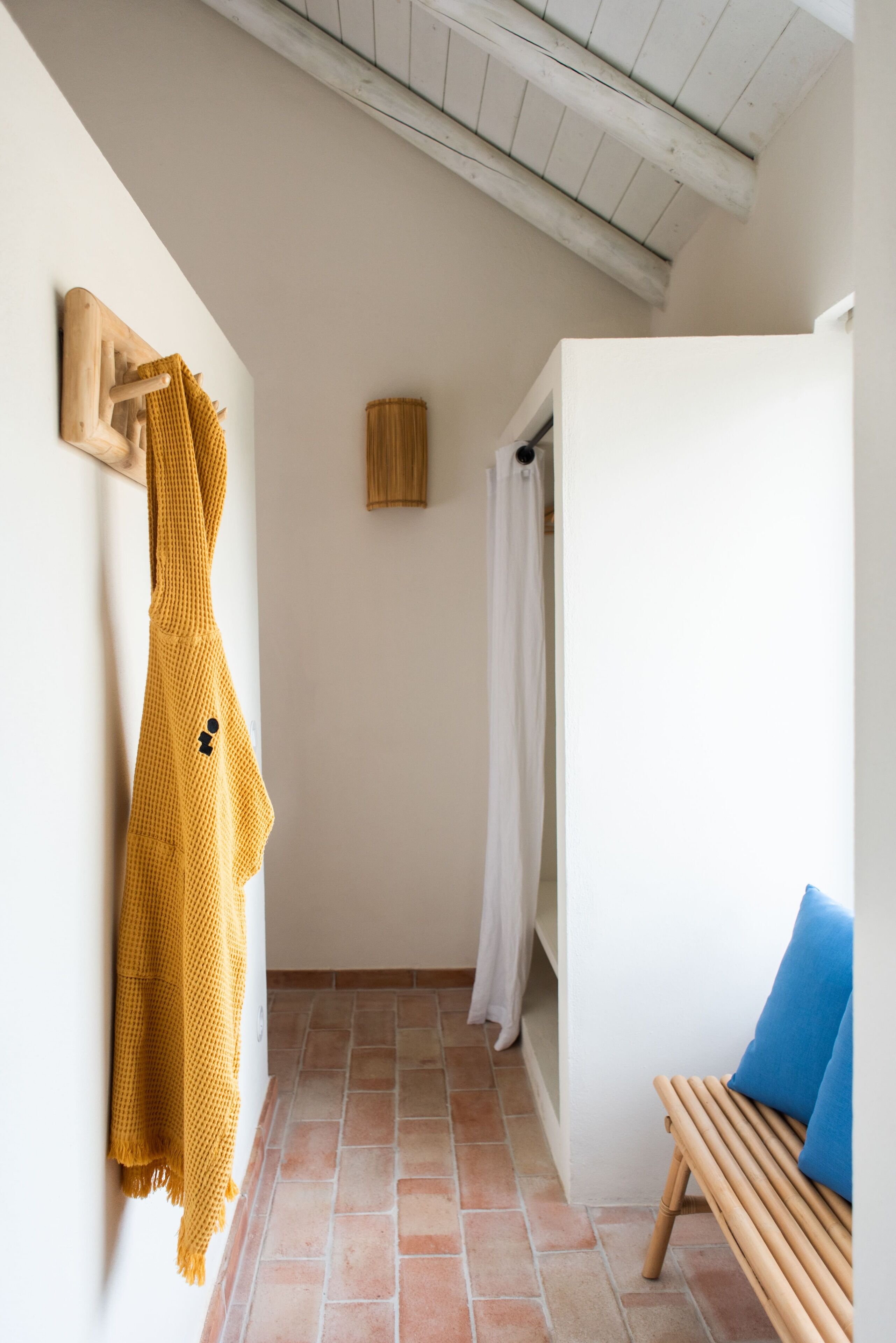 Un pasillo estrecho con un banco de madera, un objeto amarillo tejido colgado en la pared y una puerta de madera.