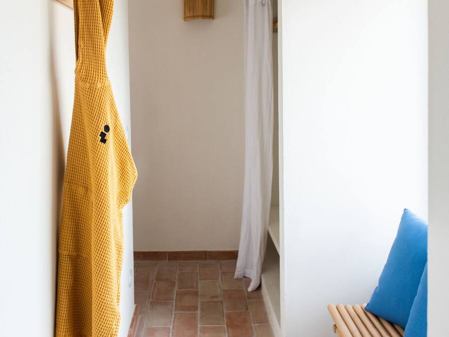 Un pasillo estrecho con un banco de madera, un objeto amarillo tejido colgado en la pared y una puerta de madera.