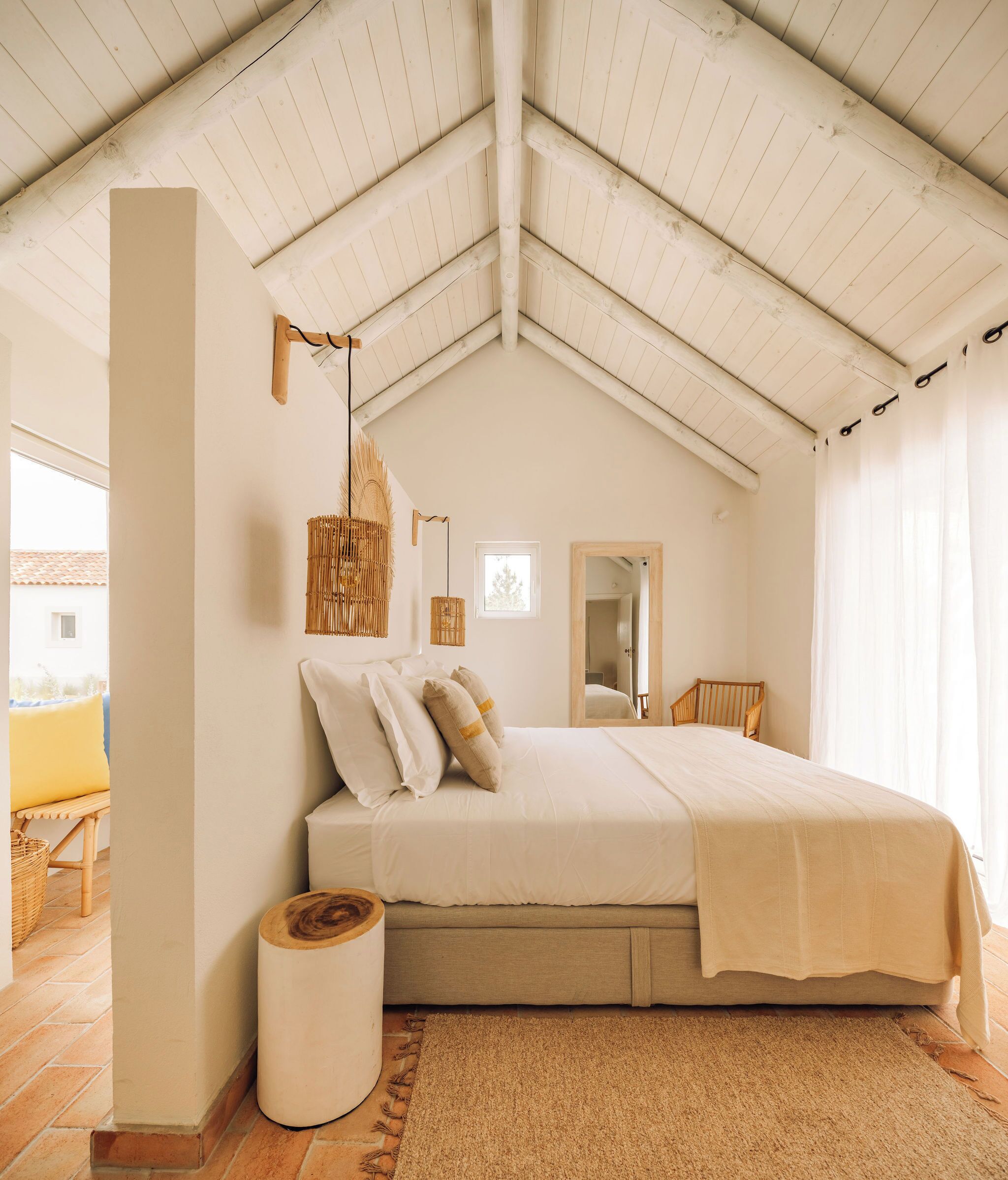 Een slaapkamer met hoog plafond, een bed met wit beddengoed, een houten vloer, een geweven vloerkleed en een groot raam.