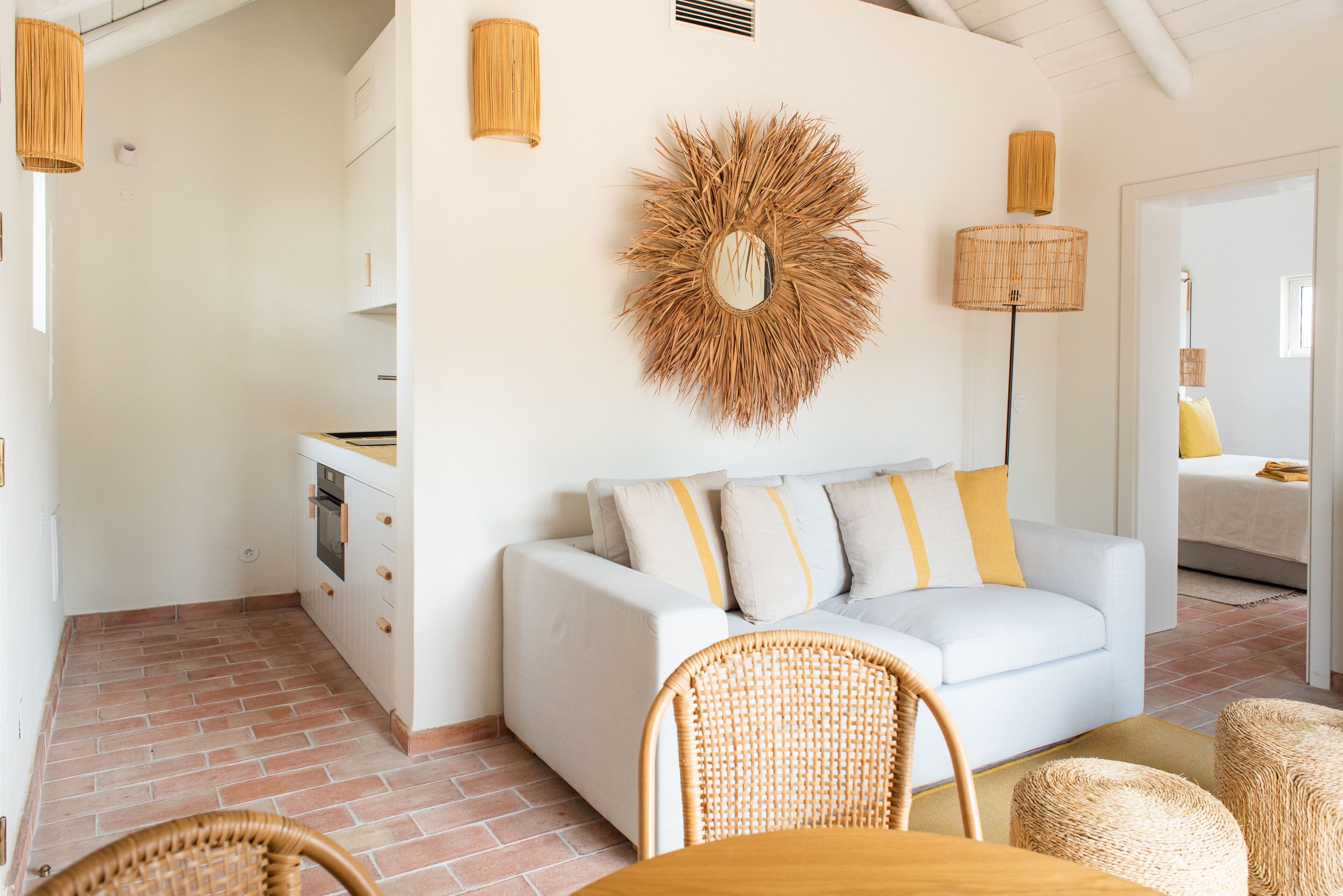 A modern kitchen with white cabinets, a white sofa with yellow and white cushions, a wicker chair, and a woven wall decoration.