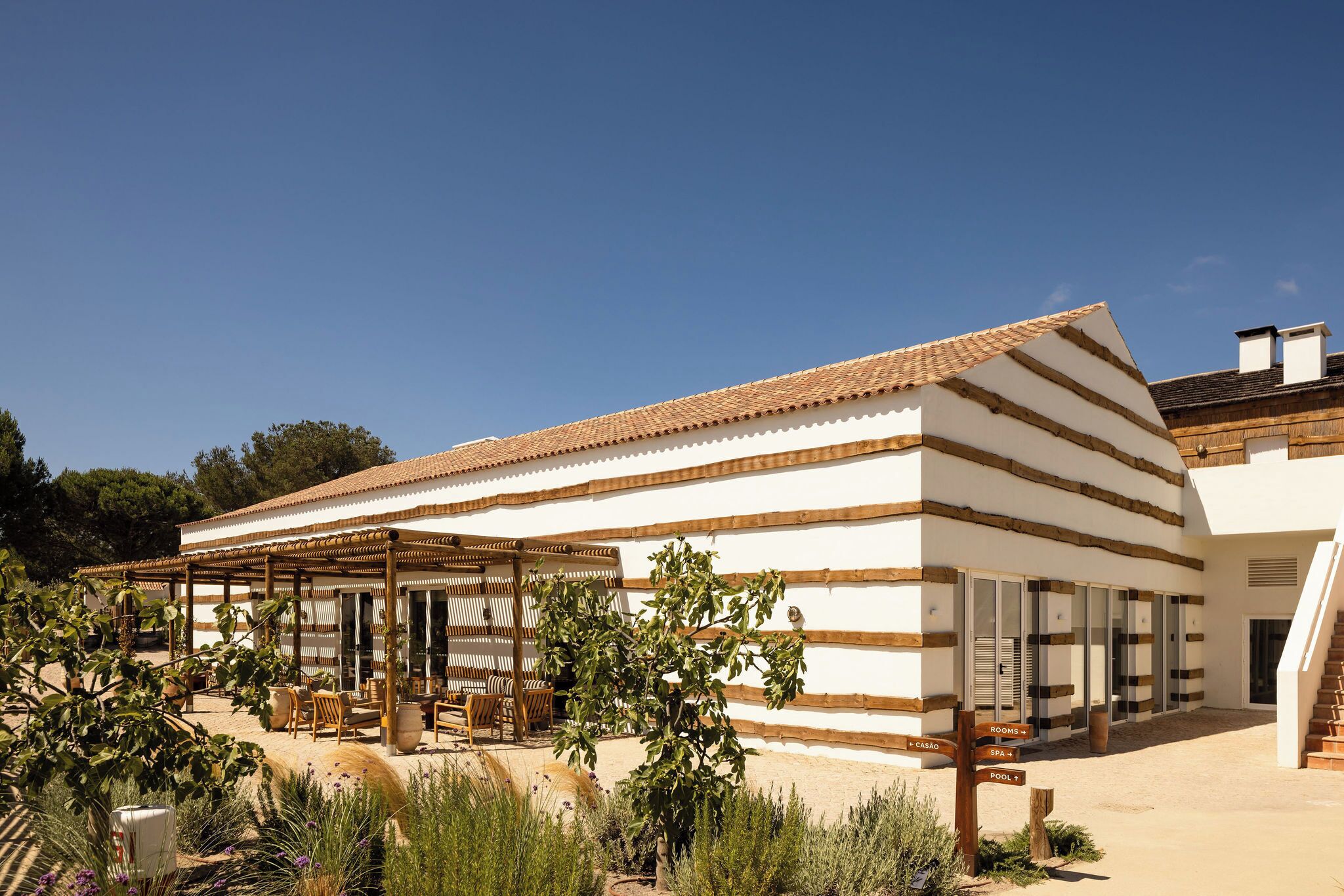A building with a tiled roof, a covered patio area with seating, and a sign indicating rooms and a pool.