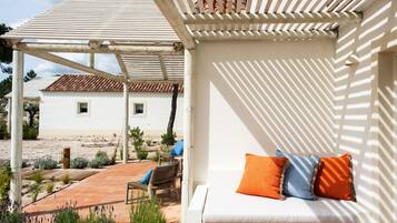 A white outdoor seating area with a pergola, a white building in the background, and a patio with plants.