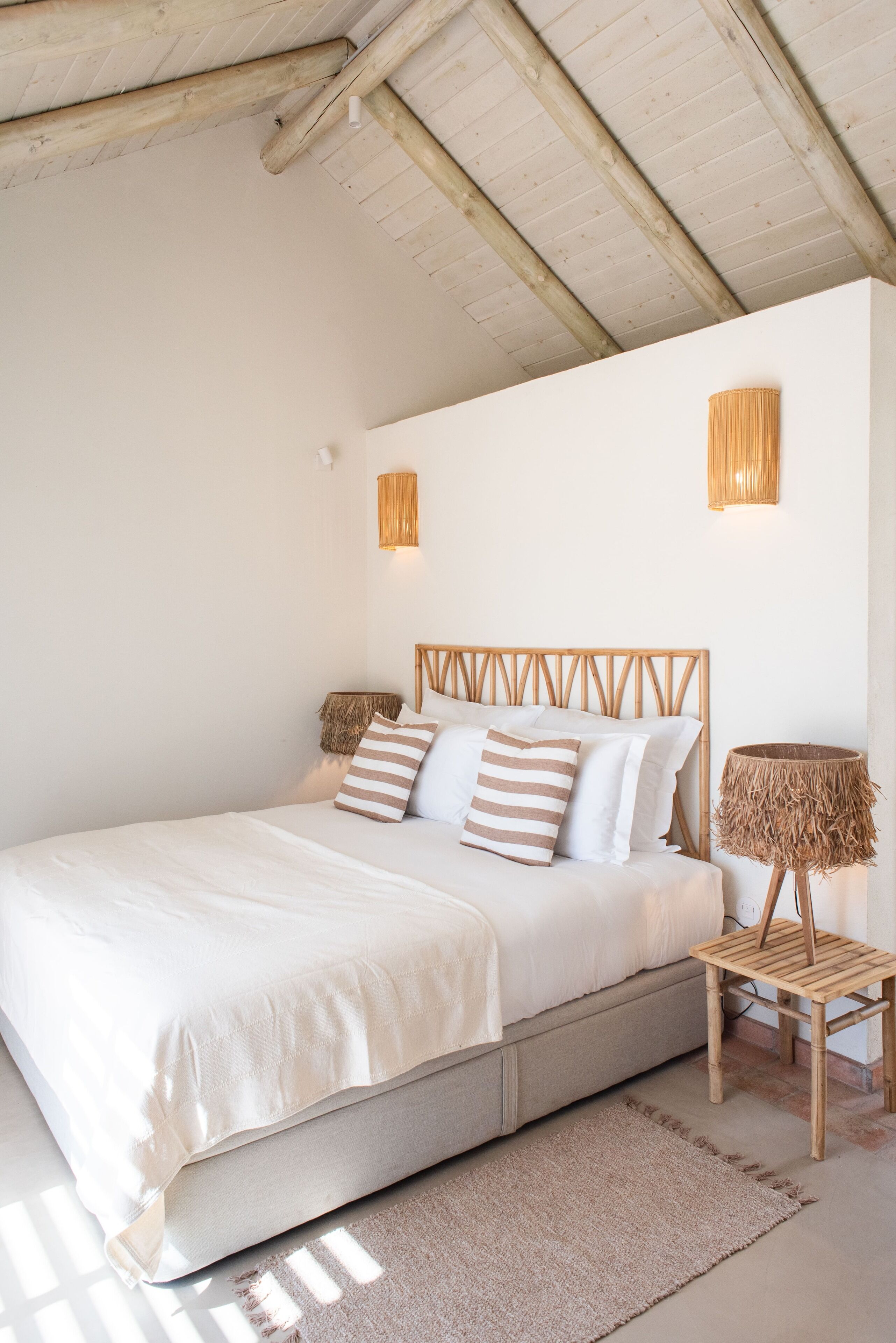 Un dormitorio con cabecera de madera, cama con ropa de cama blanca, dos lámparas fijadas a la pared y una mesita de noche tejida.