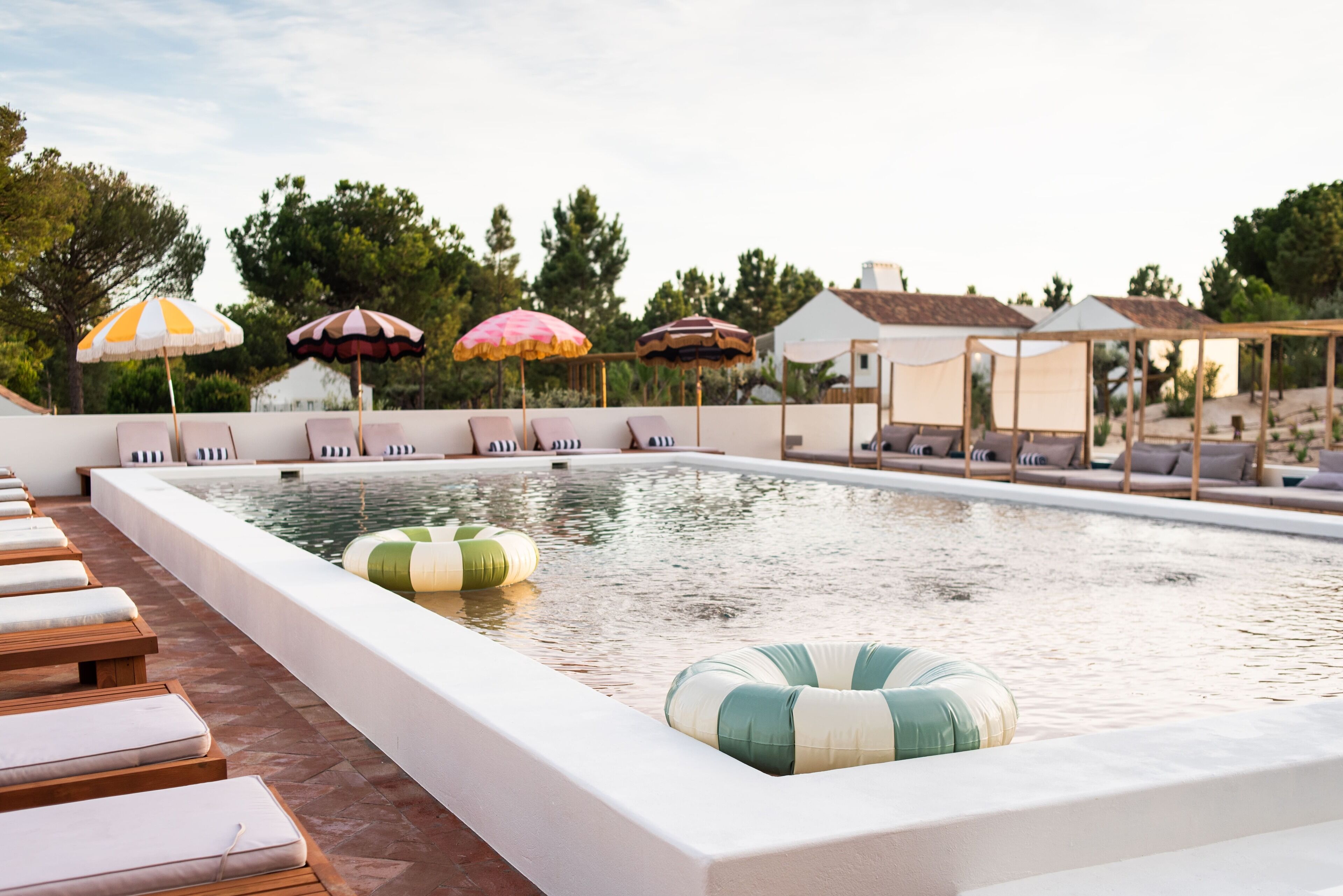Una piscina con sillas de descanso y sombrillas, rodeada de árboles y un edificio.