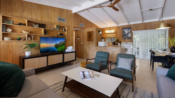 A modern living room with a wooden panel wall, a flat-screen TV, a dining area with a table and chairs, and a ceiling fan.