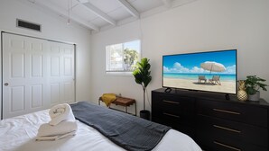 A bedroom with a bed, a TV displaying a beach scene, a bench, a potted plant, and a window with a view of the outdoors.