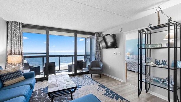 A modern living room with a large window overlooking the ocean, a flat-screen TV, a black bookshelf, and a cozy seating area with a blue sofa and a patterned rug. - Golden Sands 1517 by Seaside Vacations & Sales (Ocean City)