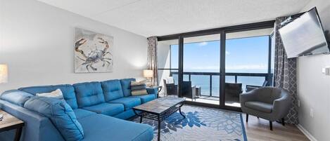 A modern living room with a blue sectional sofa, a coffee table, and a large window offering a view of the sea.