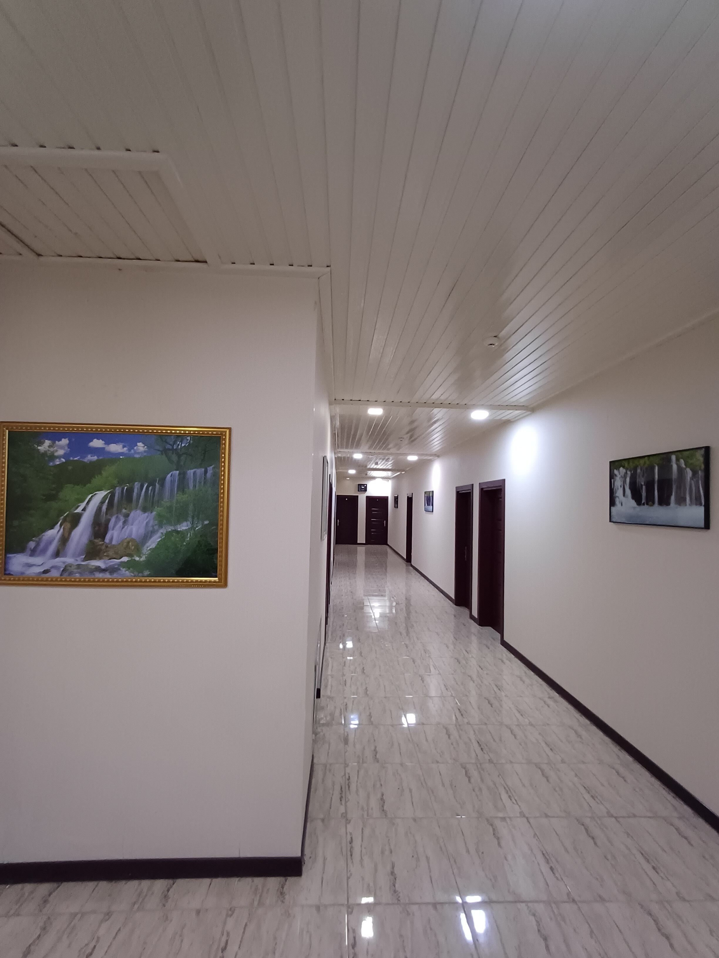 Um corredor com piso de mármore, paredes brancas e teto com painéis de madeira. Há portas à direita e um quadro emoldurado à esquerda.