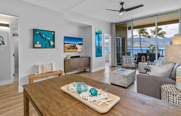 A spacious living area with a wooden dining table, a sofa, and a balcony with a view of the sea. - Upscale Oceanfront Penthouse Vistas (Kihei)