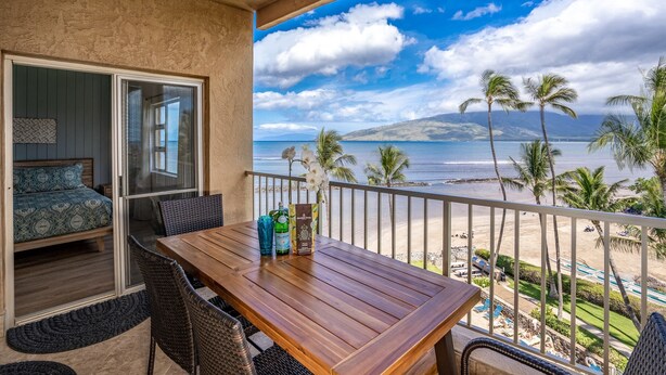A balcony with a wooden table, chairs, and a view of the beach and ocean.