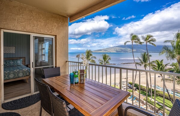 A balcony with a wooden table, chairs, and a view of the beach and ocean.