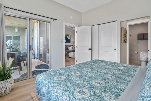 A bedroom with a patterned bedspread, a sliding glass door leading to a balcony, and a view of a dining area. - Upscale Oceanfront Penthouse Vistas (Kihei)