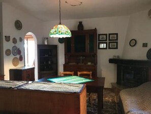 Un coin repas avec une table et des chaises en bois, un lustre suspendu en vitrail, une cheminée et un mur décoré d’assiettes et de tableaux encadrés.
