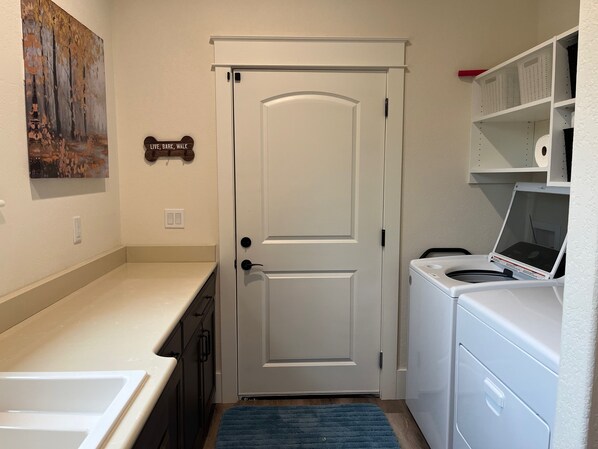 A laundry room with a white door, a washing machine, and a dryer. There is a blue rug on the floor and a painting on the wall. - The Cottage at Casa del Arbol (Tellico Village) (Loudon)