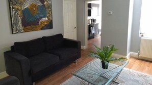 Een woonkamer met een zwarte bank, een glazen salontafel, een potplant, een wandklok en een keuken die zichtbaar is door een open deur.