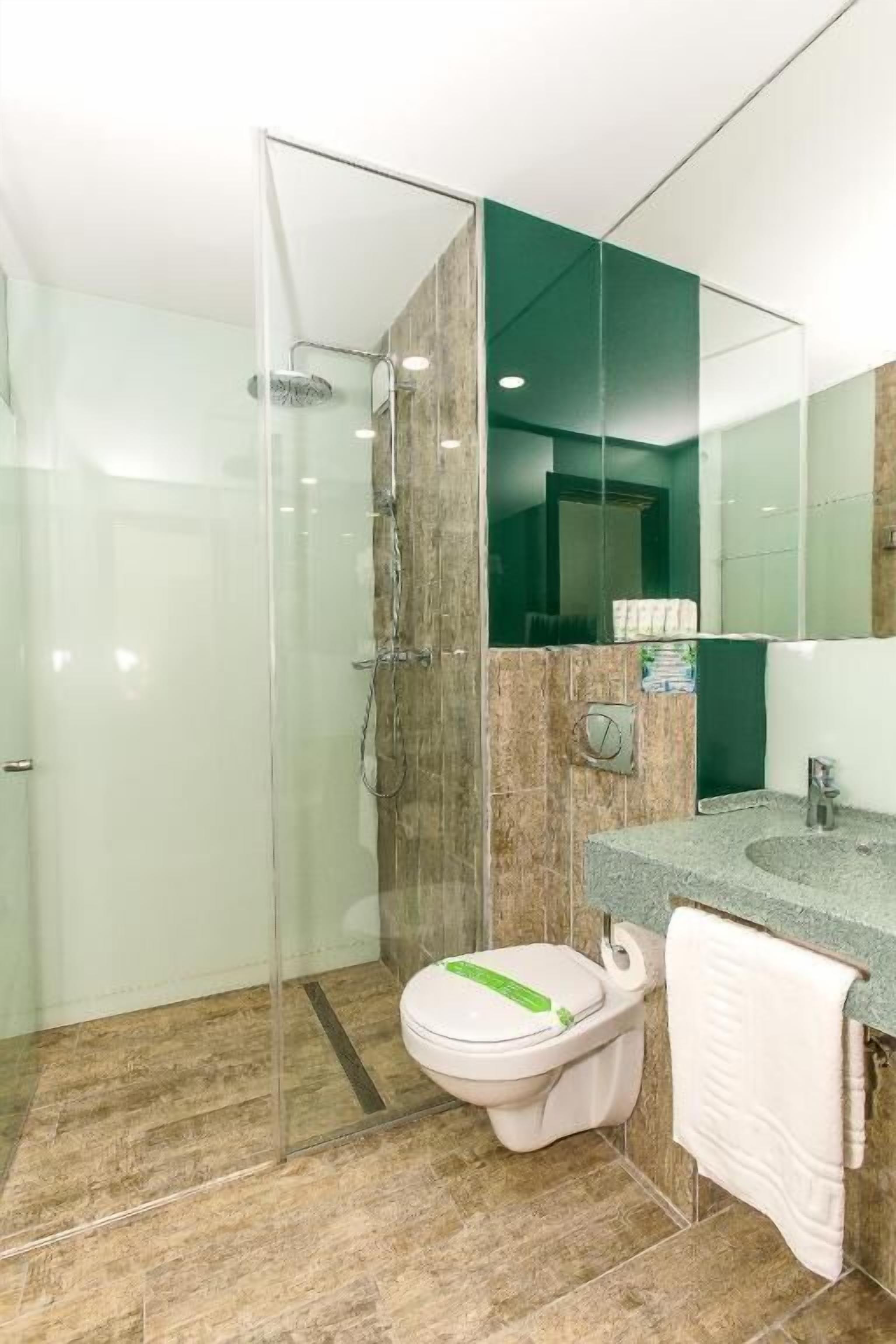 Une salle de bain moderne avec une douche à parois de verre, des toilettes dotées d’un porte-papier toilette vert et un lavabo avec une serviette accrochée au mur.