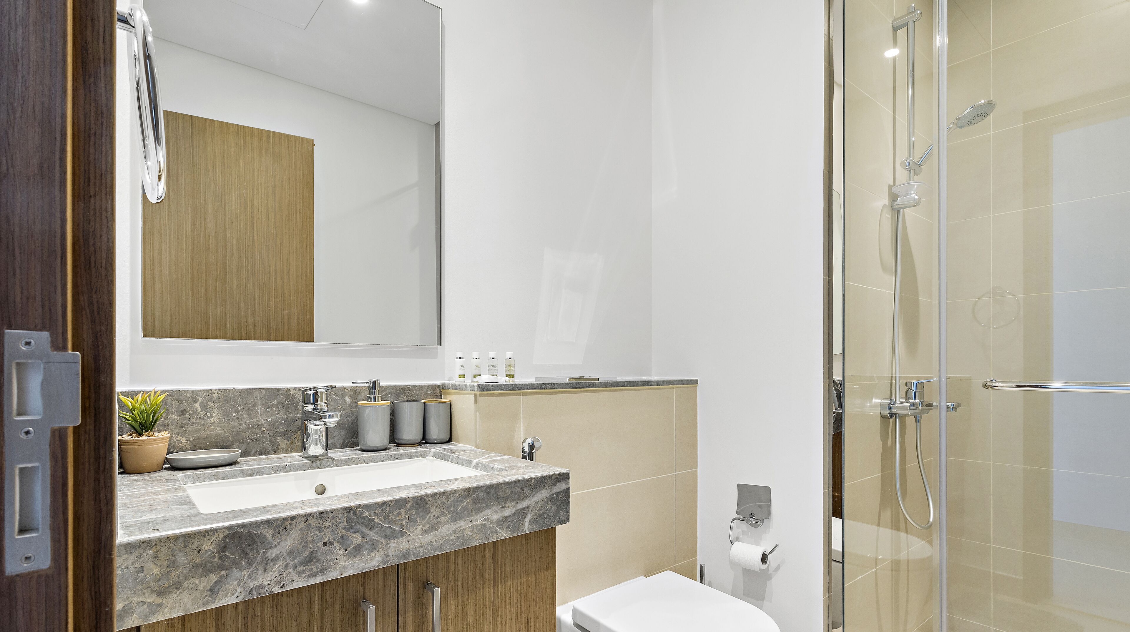 A modern bathroom with a marble sink, wooden cabinets, a glass shower enclosure, and a toilet paper holder.