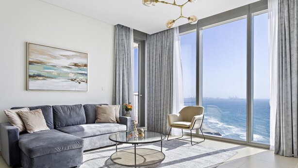 Un salon moderne avec un canapé gris, une table basse ronde et une vue sur l’océan grâce à de grandes fenêtres.