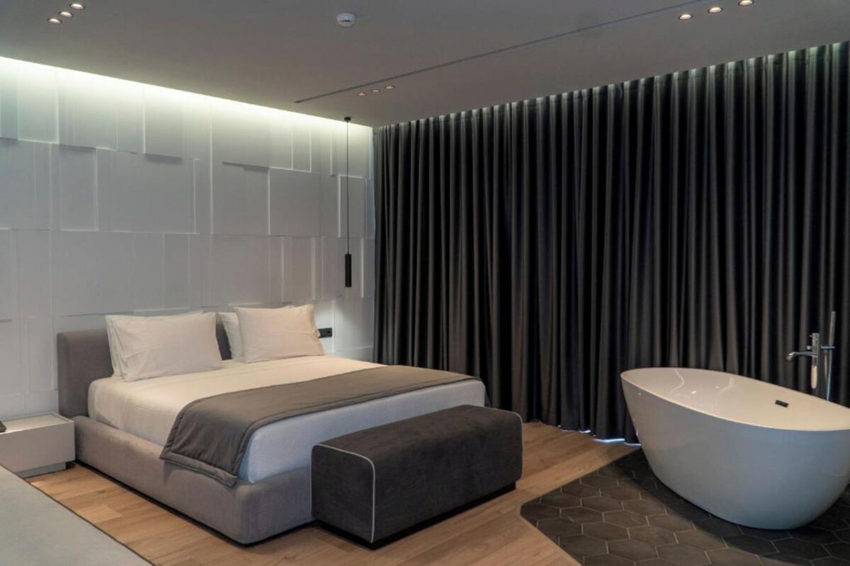 A modern bedroom with a large bed, a bedside table, a bench, and a freestanding bathtub.