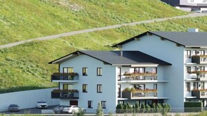 A modern multi-story building with balconies, a parking area, and a hillside backdrop.