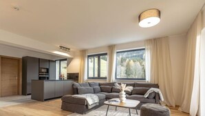 A modern living room with a sofa, ottoman, and coffee table, a kitchen area, and large windows with curtains.