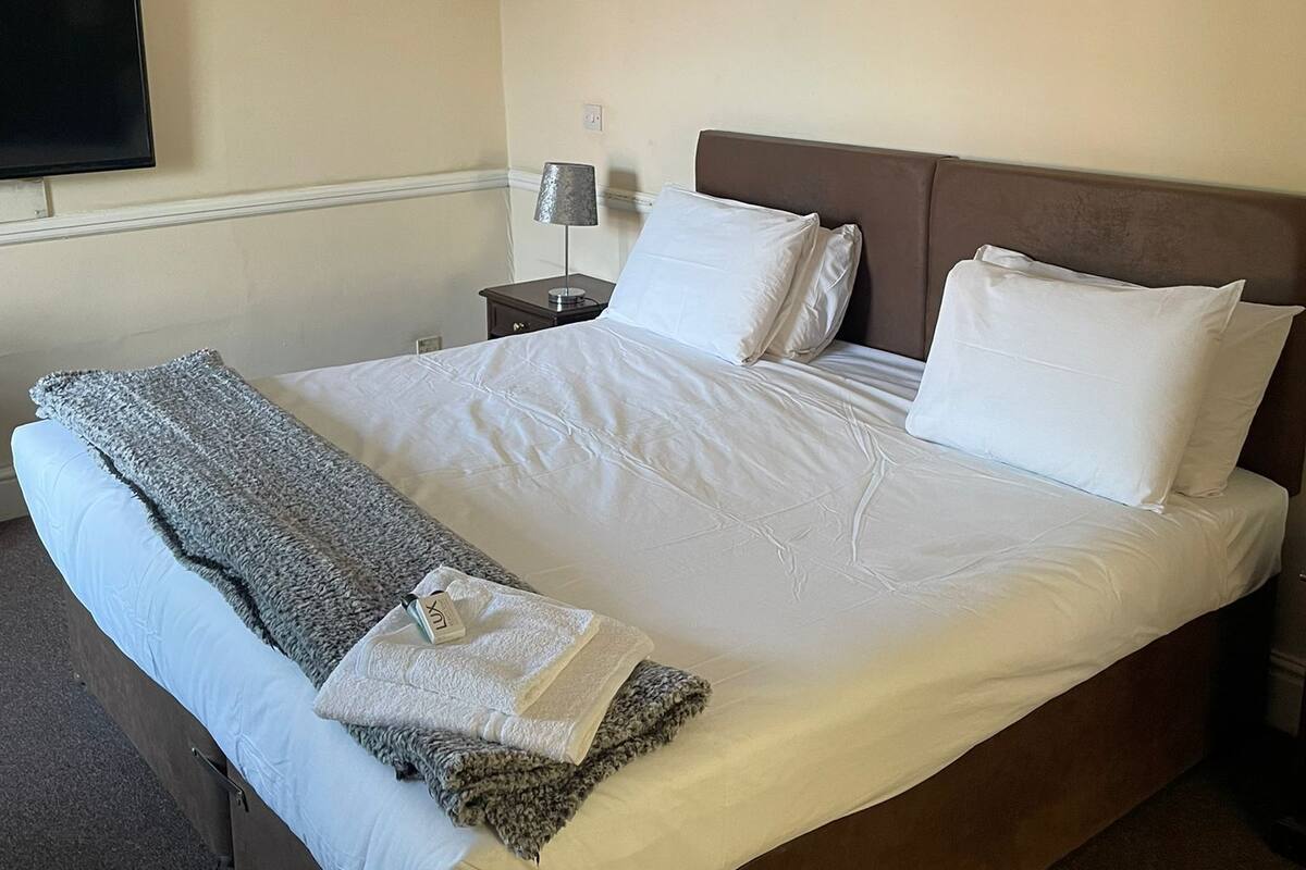 A neatly made bed with white linens and a grey blanket, a bedside lamp, and a flat-screen TV mounted on the wall.