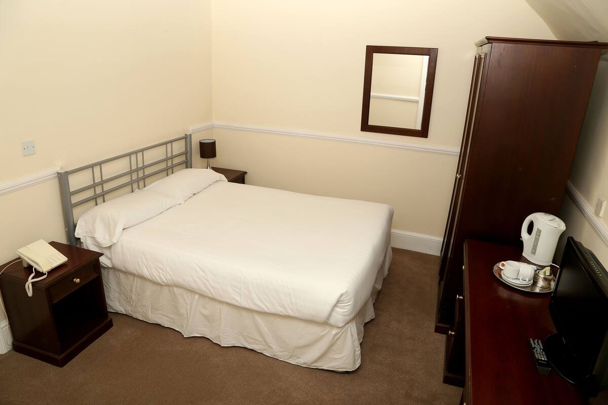 A neatly made bed with white bedding, a bedside table with a lamp, a small nightstand with a phone, a wooden dresser with a kettle and cups, and a mirror on the wall.