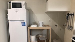 Une petite cuisine équipée avec un réfrigérateur blanc, un four à micro-ondes, une petite table en bois avec une bouilloire et un panier, ainsi qu’une poubelle.