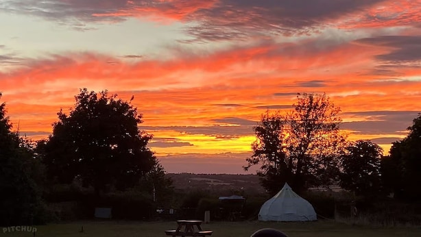 A tent and picnic table in a grassy field with a sunset sky. - Woodlands Basic Bell Tent 2 (Grantham)