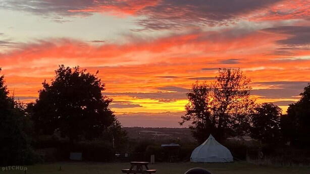 Un camping con una tienda de campaña y una mesa de picnic, rodeado de árboles y un cielo al atardecer.