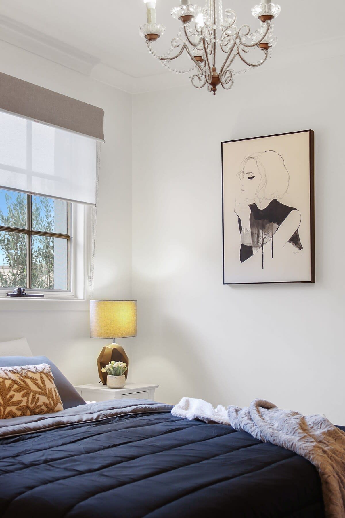 A bedroom with a bed, a chandelier, a window with blinds, a bedside table with a lamp, and a framed artwork on the wall.