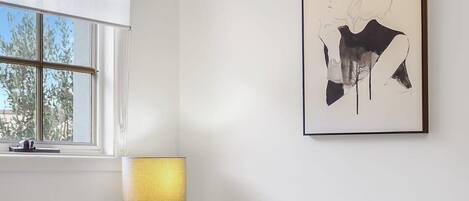 A bedroom with a bed, a chandelier, a window with blinds, a bedside table with a lamp, and a framed artwork on the wall.
