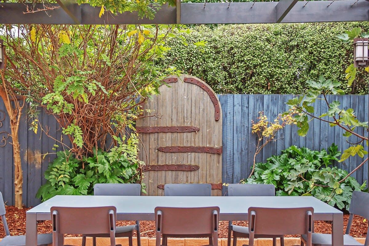 An outdoor patio with a rustic wooden gate, a table with chairs, and a hedge in the background.