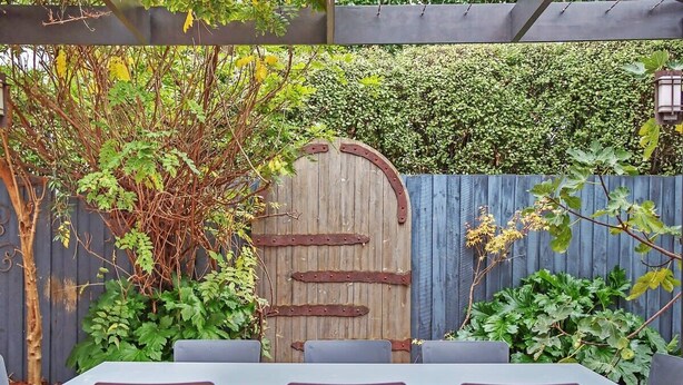 Un patio al aire libre con un portón de madera rústica, una mesa con sillas y un seto al fondo.