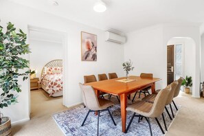 A dining area with a wooden table and chairs, a bed with floral bedding, and a potted plant.