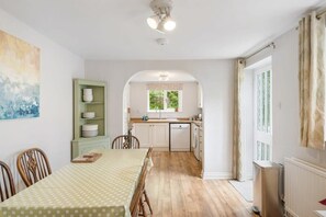 A dining area with a table set for four, a green shelving unit with dishes, and a view into the kitchen through an archway.
