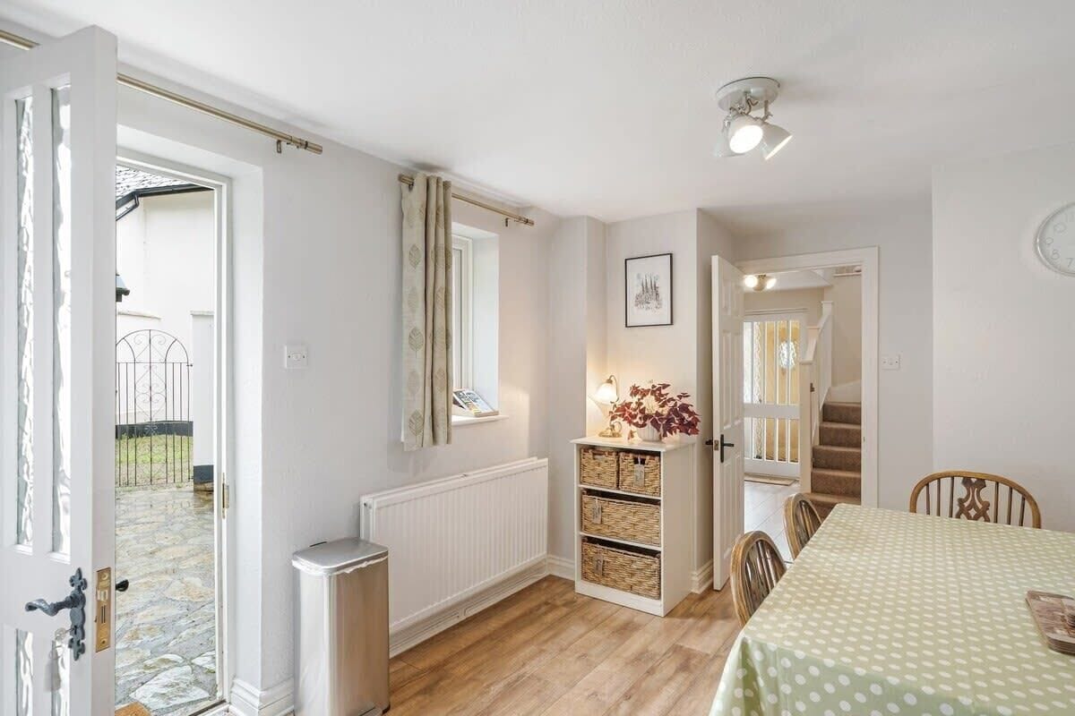 Une pièce avec un plancher en bois, une table à manger recouverte d’une nappe verte, un radiateur, une étagère avec des paniers et un escalier menant à une autre pièce.